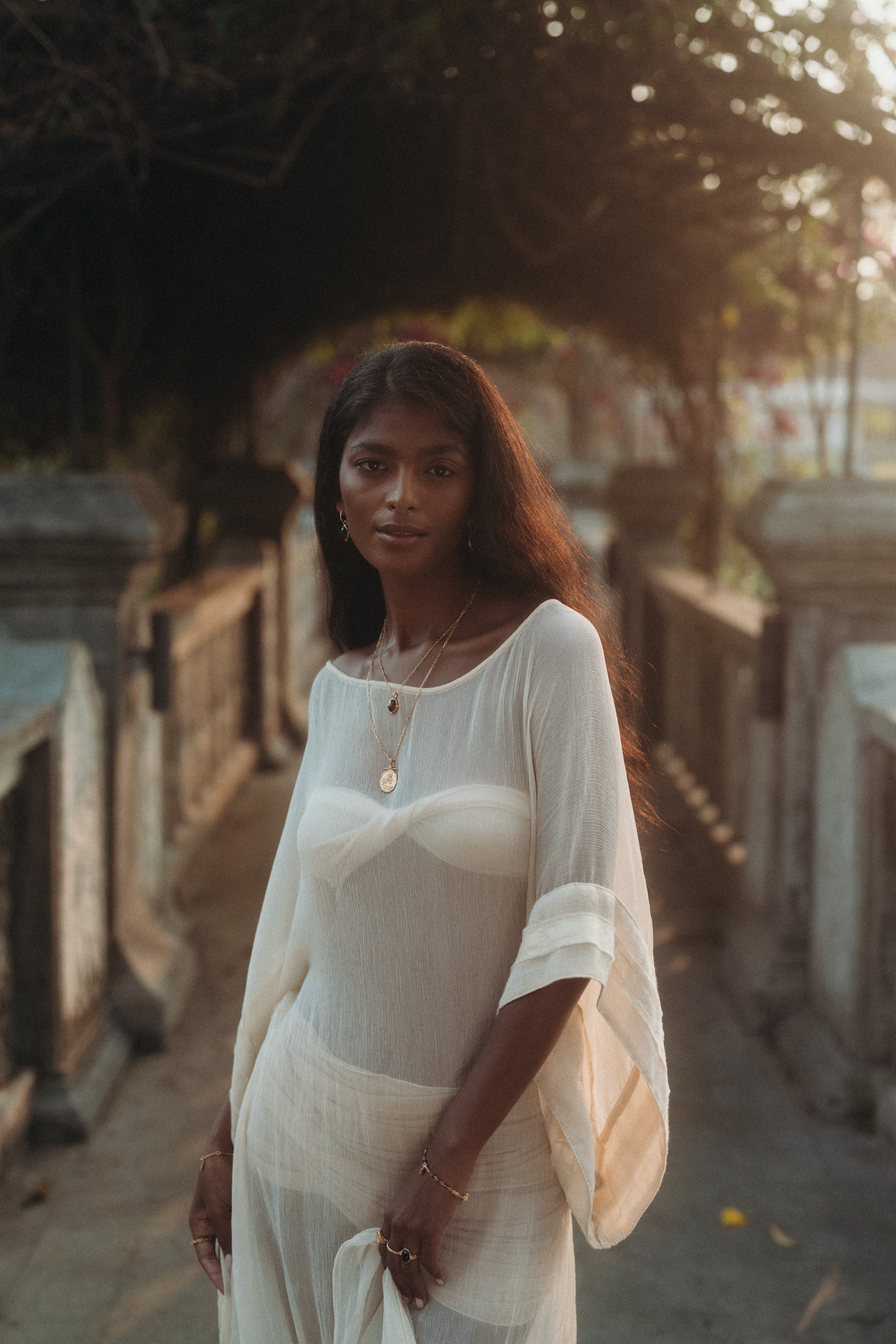Woman in a sheer, flowing Sundari Kaftan from Bird of Paradise, made of natural silk chiffon, standing in golden sunset light. This luxury sustainable fashion piece embodies Bali boho clothing and slow fashion elegance.