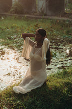 Woman kneeling by a lotus pond at sunset, wearing the Sundari Kaftan from Bird of Paradise. This sheer silk chiffon dress embodies luxury sustainable fashion, bohemian elegance, and the essence of Bali boho clothing.