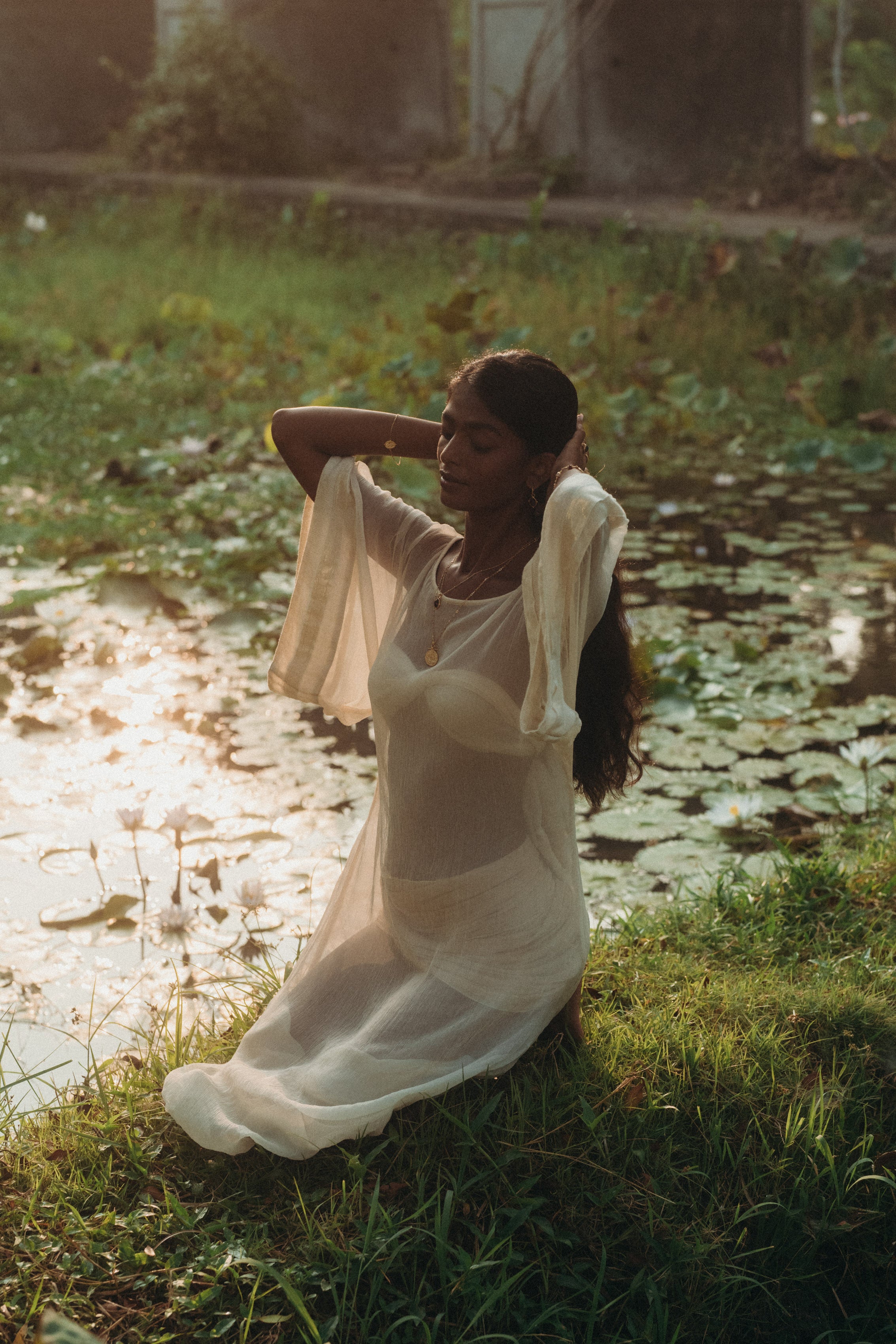 Woman kneeling by a lotus pond at sunset, wearing the Sundari Kaftan from Bird of Paradise. This sheer silk chiffon dress embodies luxury sustainable fashion, bohemian elegance, and the essence of Bali boho clothing.