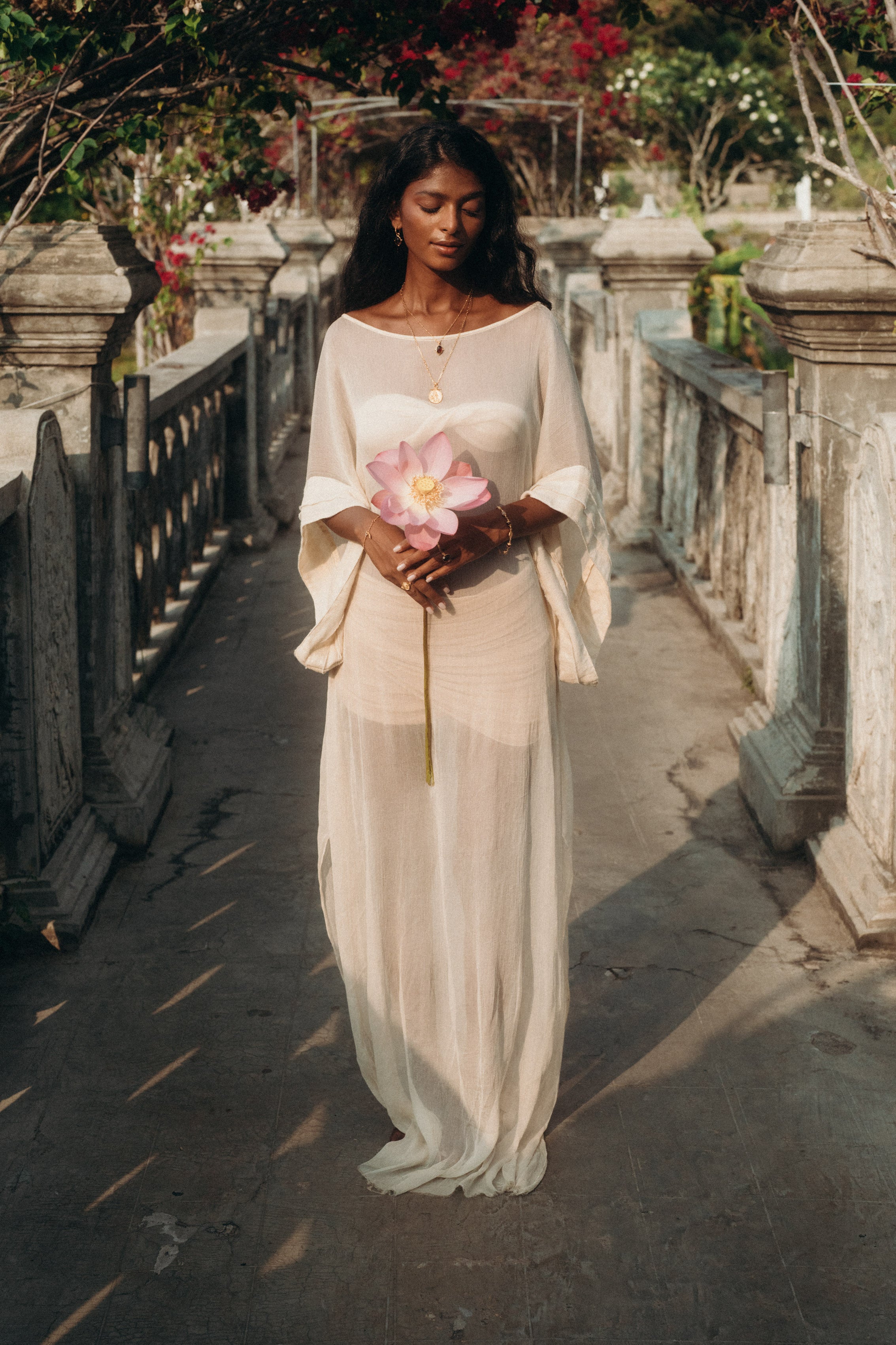 Woman standing gracefully in the Sundari Kaftan from Bird of Paradise, a sheer silk chiffon dress that embodies bohemian elegance and mindful fashion. Holding a pink lotus flower, she reflects the essence of Bali boho clothing and luxury sustainable fashion.
