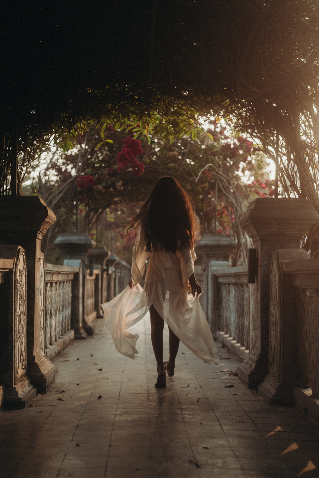 Woman walking barefoot through an ornate pathway at sunset, wearing the flowing Sundari Kaftan from Bird of Paradise. This sheer silk chiffon dress embodies bohemian elegance, slow fashion, and mindful luxury in Bali boho clothing.
