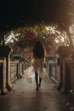 Woman walking barefoot through an ornate pathway at sunset, wearing the flowing Sundari Kaftan from Bird of Paradise. This sheer silk chiffon dress embodies bohemian elegance, slow fashion, and mindful luxury in Bali boho clothing.